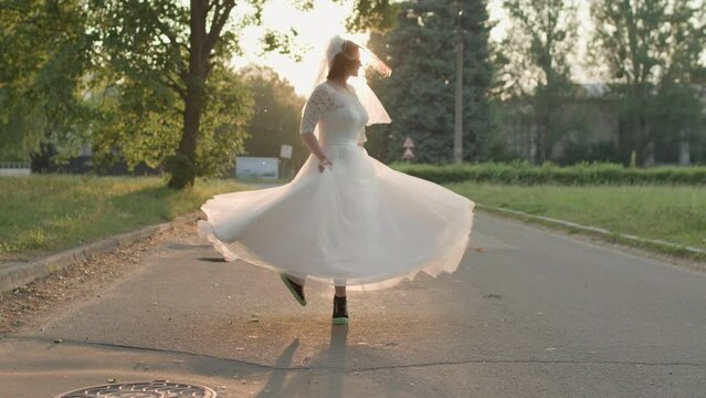 Bride in white wedding dress and snickers spinning around on the road at sunrise. Slow motion, handheld camera.