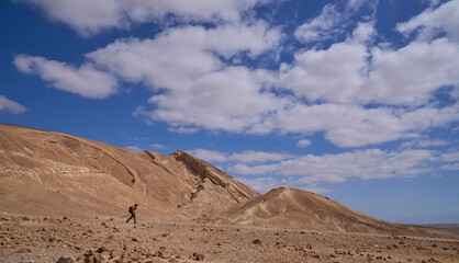 Male hiker on a trail in Negev desert. Orange and yellow colors of the mountains rocky landscape. Almost vertical wall of the Small Crater at the background. Popular hiking in park Hamakhtesh Hakatan.