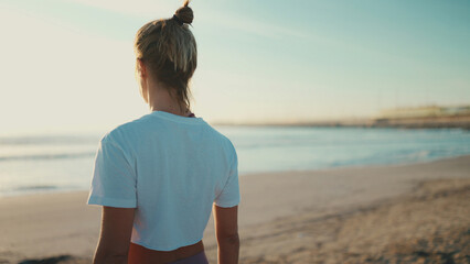 Back view of blond woman enjoying good day after yoga practice on the beach. Sporty girl standing...