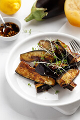 Grilled fried eggplant sticks with soy sauce and honey, garnished with microgreens, on white plate and white background, delicious vegetarian dish.