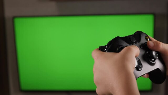 Close-up of a hand with a joystick in front of a green screen TV, a teenager is playing video games. Place for your advertisement. Chroma key.