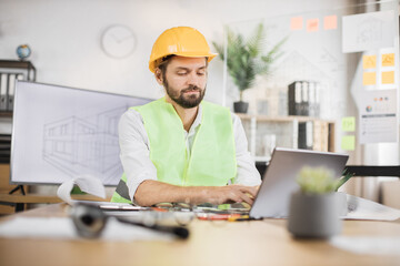 Competent engineer, designer or architect leading working conference at office center. Attractive young bearded man wearing uniform and protective helmet working on laptop with blueprint.