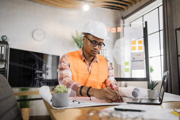 Competent African American engineer, designer or architect leading working conference at office center. Attractive young man wearing uniform and protective helmet working on laptop with blueprint.