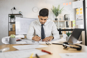 Competent arhitect indian male sitting on desk indoors using pen and ruler for project drawings. Positiv hindu bearded young man wearing white shirt and black tie.