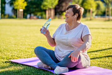 Relaxed senior plus size woman with earphones sitting on yoga mat on green grass outdoors resting...
