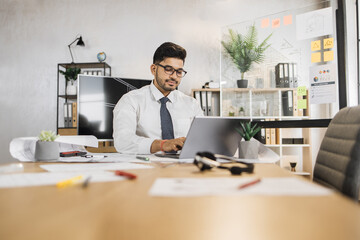 Professional Arab Muslim male architect using laptop computer, sitting on the desk while working on construction plan, using sketches at his workplace.