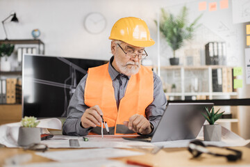 Qualified mature architect in reflective vest uniform and hardhat and glasses checking construction project with blueprint and laptop sitting on table. Bearded senior man controlling working process.