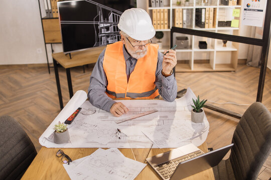 Competent architect senior male sitting on desk indoors with digital laptop using pen for project drawings. Positiv bearded mature man wearing orange reflective vest, and construction hard hat.