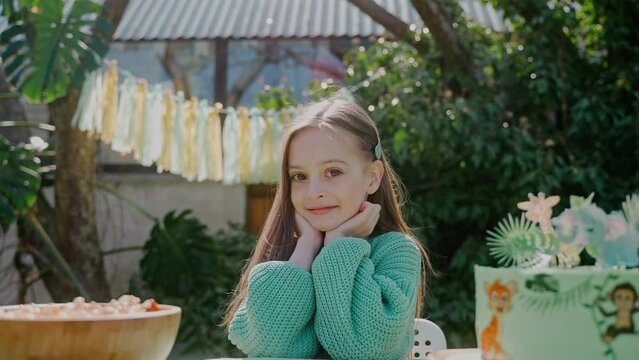 Birthday girl looks at camera sitting at the backyard party on a sunny day Green-themed birthday party. European little girl wearing a birthday hat and smiling at camera. High quality 4k footage