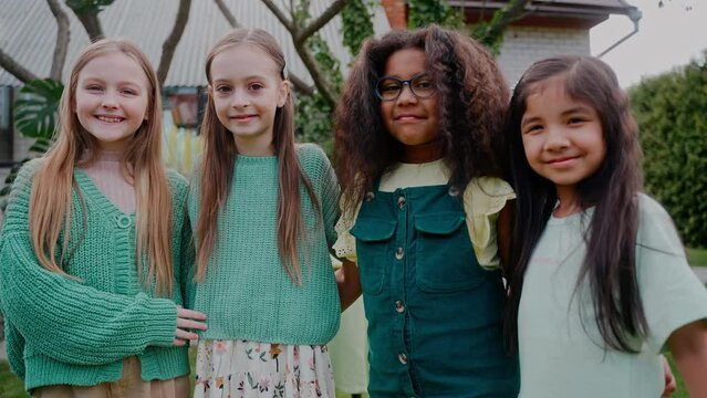 Four multinational little girls look at the camera hugging at the backyard. Green-themed birthday party. fFour girlfriends are standing in an embrace and looking at the camera. High quality 4k footage