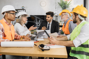 Group of five multiracial people architects sitting at the desk in modern office and discussing common project. Concept of cooperation, construction and teamwork.