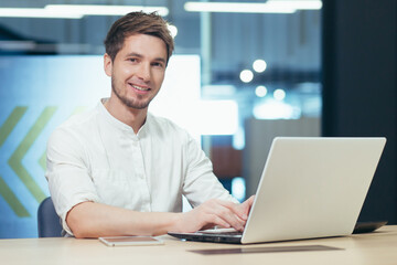Working environment. Portrait of a young handsome male businessman. Sitting at a desk in the office, working on a laptop, tablet, cell phone. Gains, works. He looks at the camera, smiles