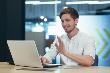 Working video call in the office. Young male businessman, freelancer sitting at a desk in the office on a laptop, talking through the camera on a video call, explaining something, waving his hands.
