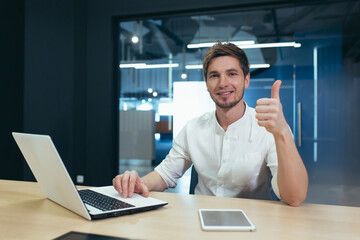 Portrait of a young successful handsome businessman, freelancer, manager, IT specialist. He sits at a table in the office, shows his hand super, looks at the camera, smiles