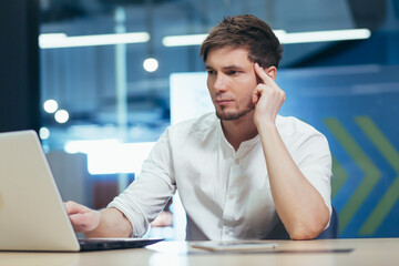 Focused and serious young man working on laptop and with documents in the office at the desk. Holds his head, He thinks