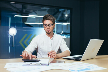 Fototapeta premium Portrait of a young handsome man, accountant, director, businessman, manager. Sitting in the office at the desk with glasses, working on a laptop, with a calculator and documents.