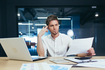 Focused and serious young man IT specialist, manager, businessman working on laptop and with documents in the office at the desk. Holds his head, he thinks