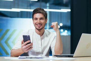 Young coassive successful man sitting in the office at his desk. Holds a mobile phone in his hand,...