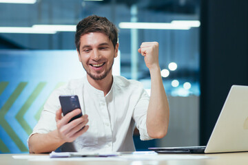 Young coassive successful man sitting in the office at his desk. Holds a mobile phone in his hand,...