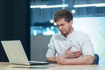 Feelings of fatigue in the workplace. Young man office worker, freelancer, IT specialist tired, holding his hand over his heart, feeling pain, discomfort. Needs rest, medical care