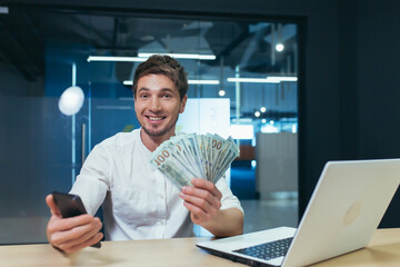 Happy and satisfied young handsome man. Businessman holding a wad of money and a mobile phone. Sits in the office at the desk, shows, brags. smiling