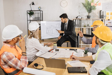 Five multiracial partners working with blueprints at office sitting near big digital screen. Professional construction team discussing arrangement of service lines at new building.