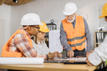 Multiracial architect people in helmets sitting at table with blueprints and modern devices. Focused workers cooperating to create new building project.