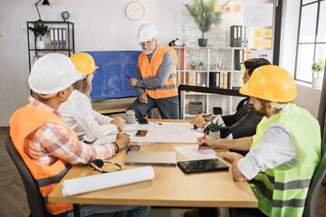 Senior male builder in hard hat showing drawings of new building on large digital screen. Group of five international partners having working meeting to discuss design and measurements of new building