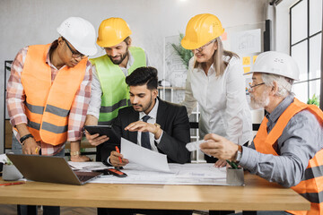 Multicultural partners working with drawings inspection during conference at modern office. Business colleagues wearing suits, uniform and protective helmet. Construction concept.