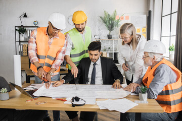 Group of five multiracial people in suits and helmets brainstorming for common project of new building. Construction team working with blueprints and modern laptop at office.