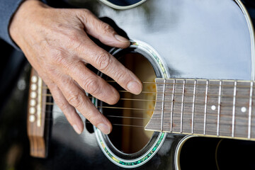 Elderly man right hand with amputated middle finger playing a black acoustic guitar - obrazy, fototapety, plakaty