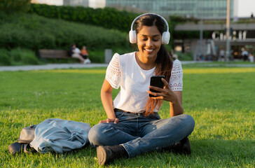 Smiling Indian woman holding mobile phone listening music in wireless headphones sitting on grass. Happy asian hipster using mobile app shopping online, reading text message in park