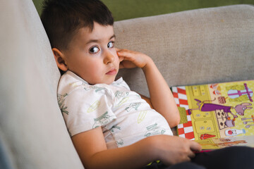 Child boy reading a book sitting on the sofa, looking at camera. Child looks at open book. Homeschool lesson.