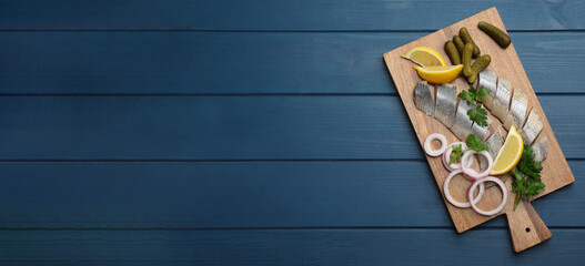 Sliced salted herring fillet served with parsley, pickles, onion rings and lemon on blue wooden table, top view with space for text. Banner design