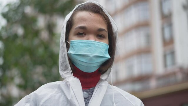Close-up portrait of confident Caucasian little woman in Covid-19 face mask looking at camera standing outdoors. Young little person posing in slow motion on city street on coronavirus pandemic