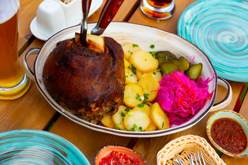 German ham hock with potato, red cabbage and sliced pickled cucumbers served on wooden table with glass of beer. - obrazy, fototapety, plakaty