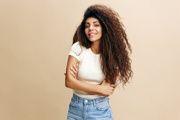 Female portrait. Happy enjoyed Latin woman with afro with folded arms smiling at camera, wearing white t-shirt, posing isolated over beige studio background. Positive casual female model laughing 