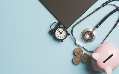 A pile of coins, an alarm clock, a stethoscope, and a piggy bank. all placed on a blue background