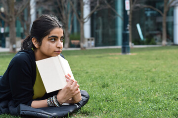 Fototapeta premium Young girl lying on green grass lawn in park reading book daydreaming
