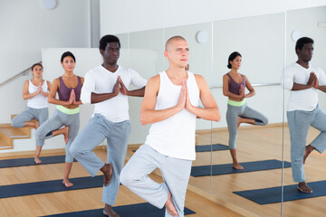 Young women and men of different nationalities exercising Hatha yoga poses in modern yoga studio.