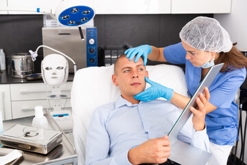 Female beautician examining face skin of young adult man before procedure at cosmetology clinic