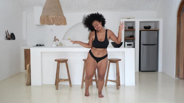 A dark-skinned, plus-size female model dances at home, oblivious to her extra mass. A full woman in underwear is body-positive, loves herself for who she is. Don't worry about body fat.