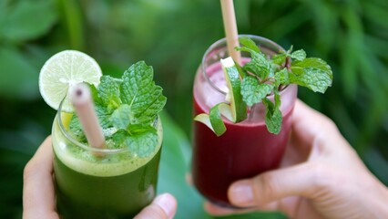 Close-up in the hands of two glasses with fresh smoothies from various fruits and berries that fill with energy and vitamins. Refreshing drinks with reusable straws and garnished with mint.