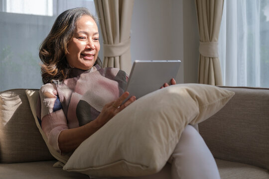 Beautiful Mature Woman Sitting In Bright Living Room And Using Digital Tablet, Surfing Internet, Checking News In The Morning.