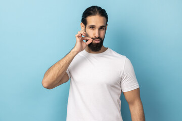I won't tell anyone. Portrait of intimidated man wearing white T-shirt zipping lips and looking at camera, covering mouth promising to keep secret. Indoor studio shot isolated on blue background.