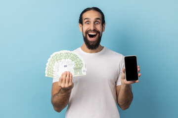 Unbelievable mobile banking application. Man wearing white T-shirt holding euro banknotes and cell phone with empty display, mock up for advertise. Indoor studio shot isolated on blue background.