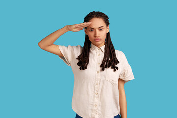 Fototapeta premium Yes sir. Portrait of responsible serious woman with black dreadlocks saluting commander, listening order with obedient expression, wearing white shirt. Indoor studio shot isolated on blue background.