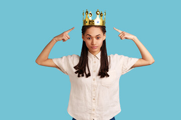 Serious independent woman with black dreadlocks points at gold crown, looking with arrogance and...