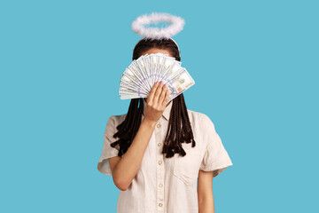 Angelic rich woman with black dreadlocks and nimbus on head hiding behind dollar banknotes, big...
