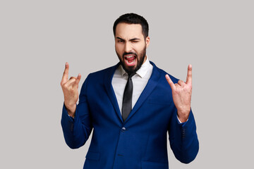 Portrait of crazy bearded man showing rock and roll sign hand gesture, screaming, having excited facial expression, wearing official style suit. Indoor studio shot isolated on gray background.